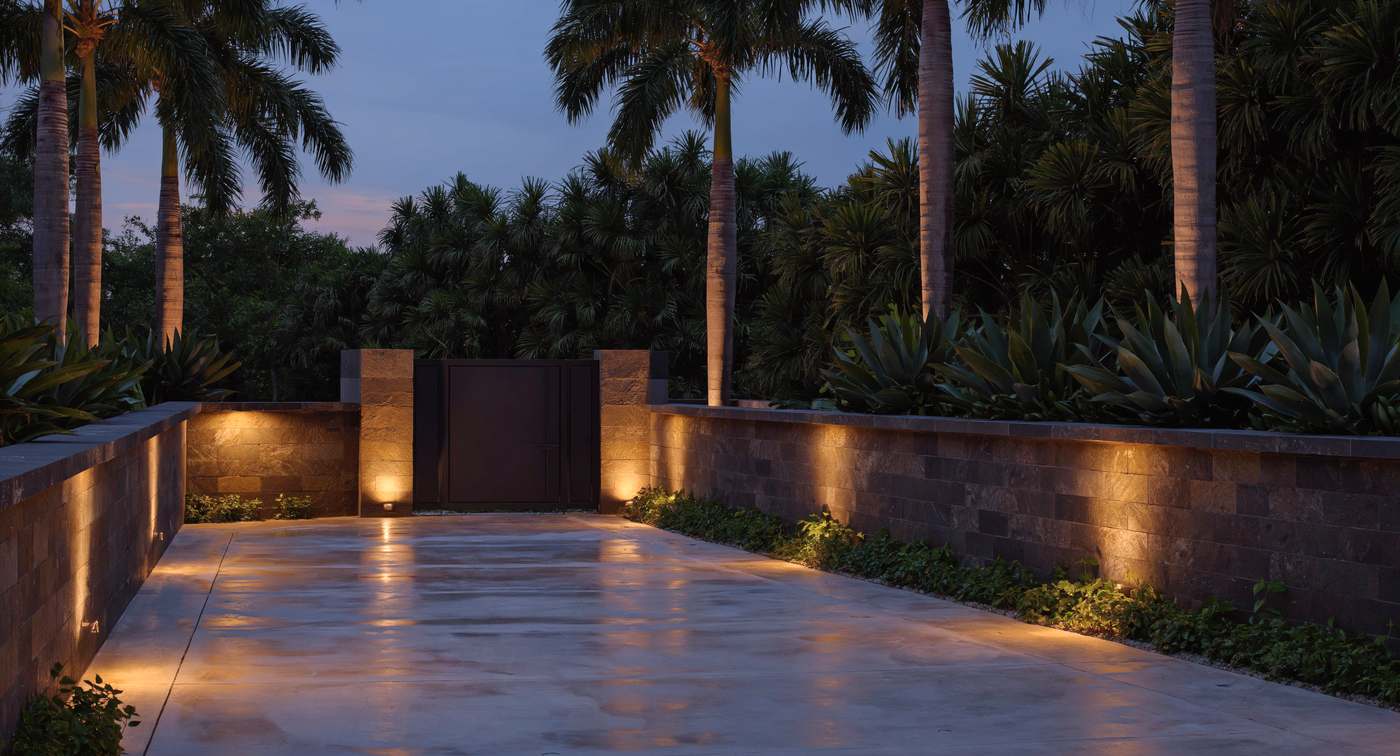 Artist's impression of an estate drive at dusk: a lit stone wall, a
                  private gate between stone piers, and palms behind.